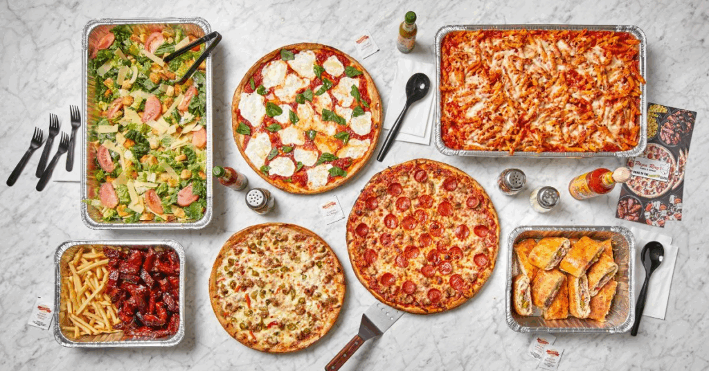 “Party table with Papa Ray’s oversized pizza, wings, and pasta trays”